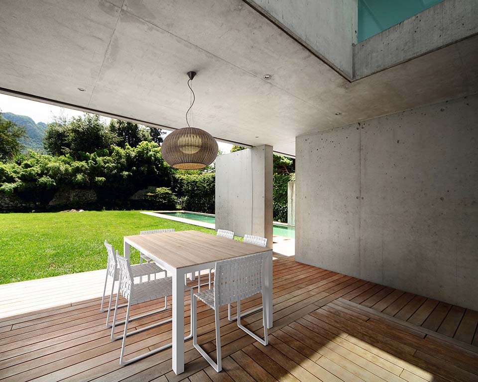 Modern covered outdoor patio with wooden deck flooring, concrete walls and ceiling, white dining table with chairs, woven pendant light, lush green lawn, and poolside contemporary garden view.