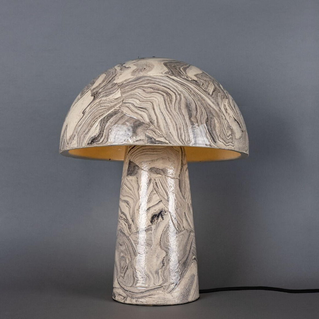 A large, elegant ceramic table lamp with a mushroom shape and a marbled black and charcoal finish, sitting on a wooden surface in a cozy, modern living space. The Amanita lamp is handmade by Mullan Lighting and features a glazed finish.