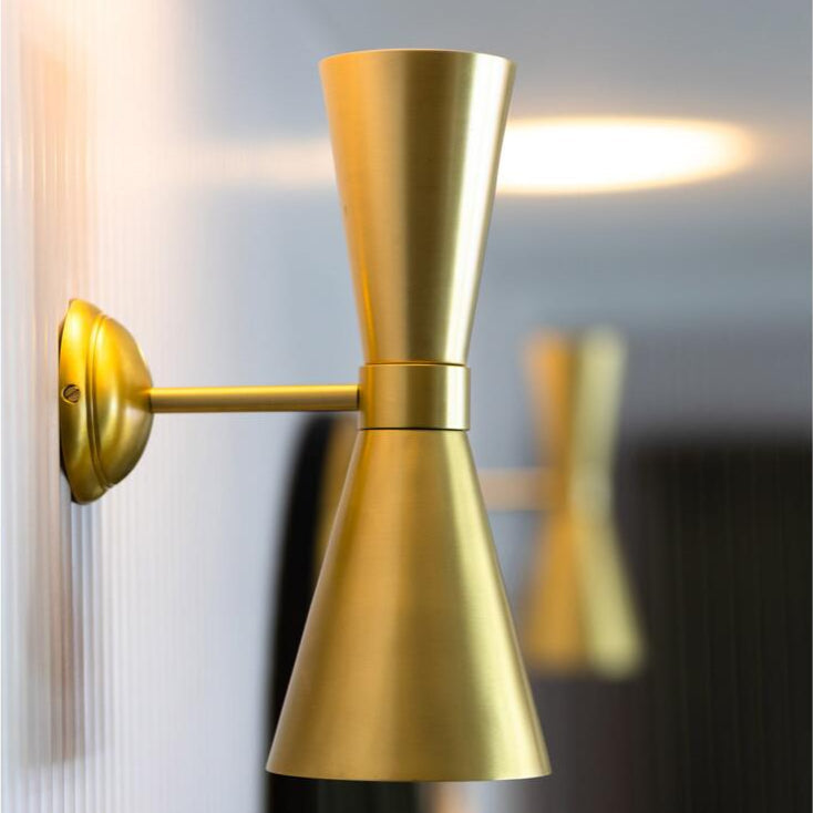 A stylish brass wall lamp with two opposing cone-shaped shades, casting light up and down on a wall. It is a sculptural piece ideal for modern and retro interiors.
