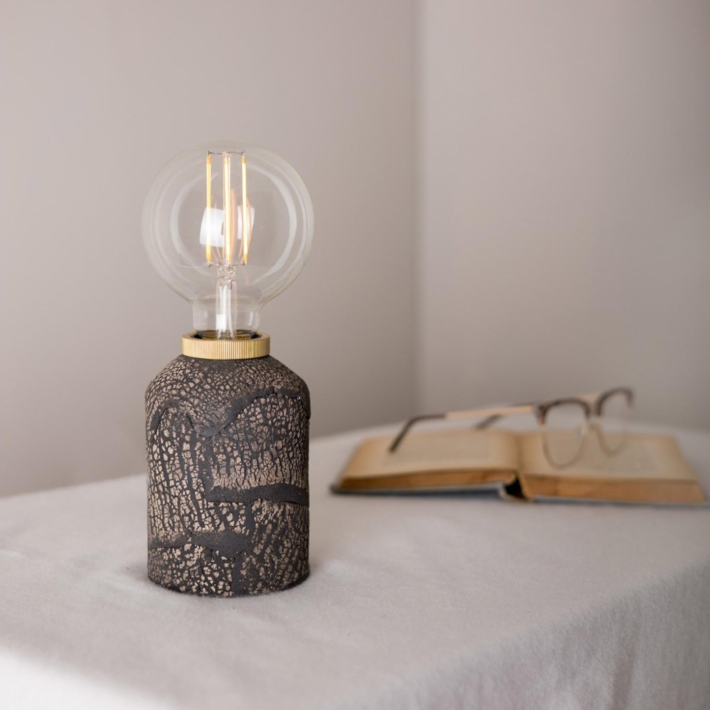 Decorative lamp with a light bulb on a surface with an open book and glasses in the background.
