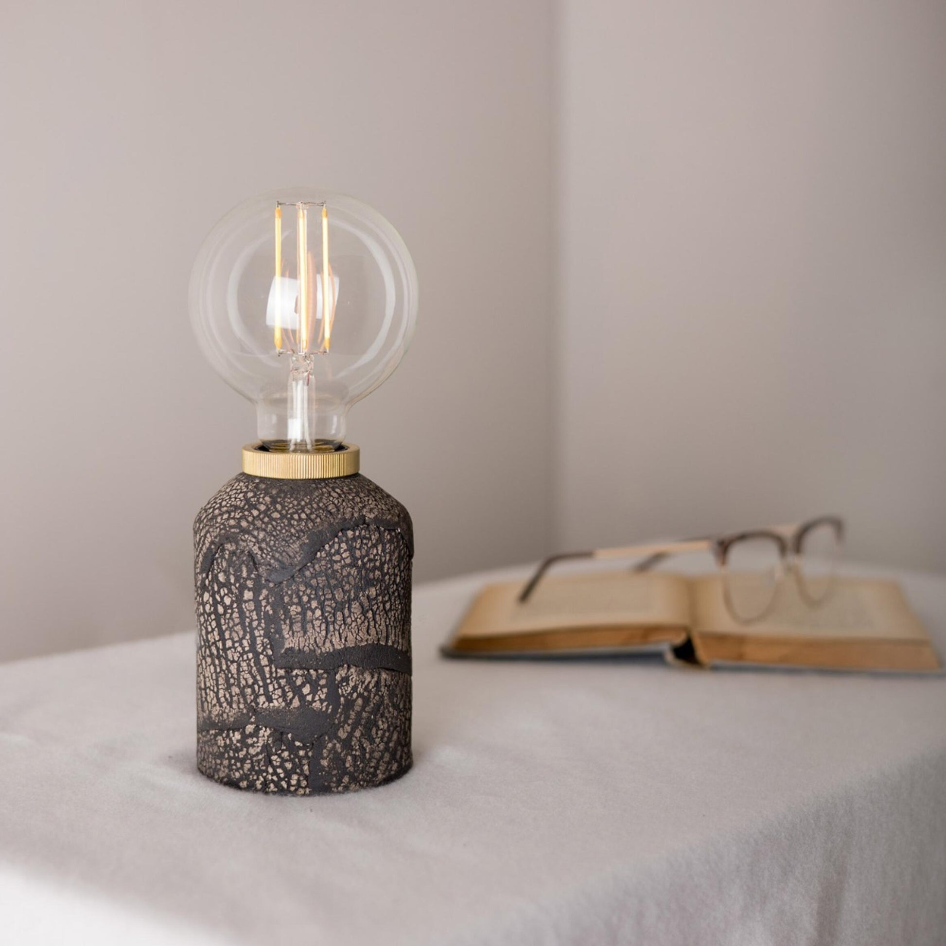 Decorative lamp with a light bulb on a surface with an open book and glasses in the background.