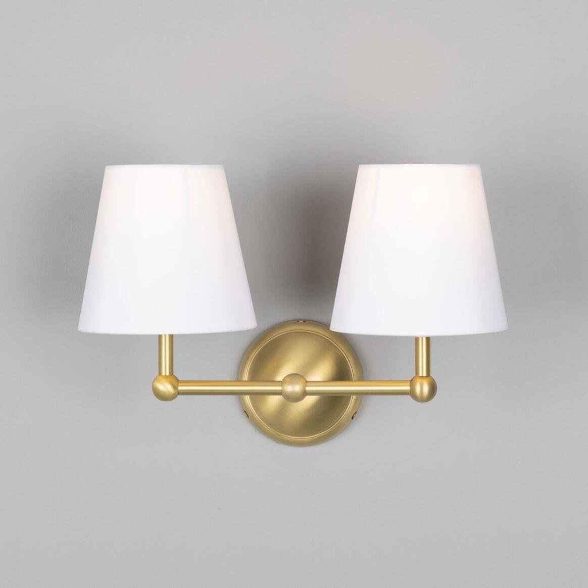 Wall-mounted light fixture with two white lampshades and gold brackets on a gray background