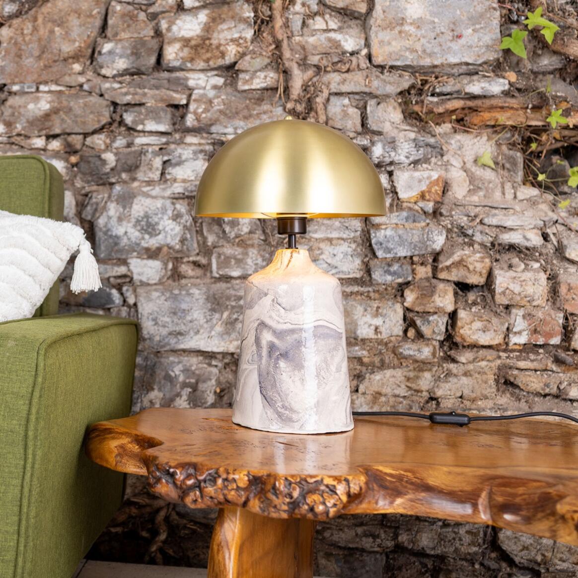 Table lamp with marble base and gold shade on a wooden table against a stone wall.
