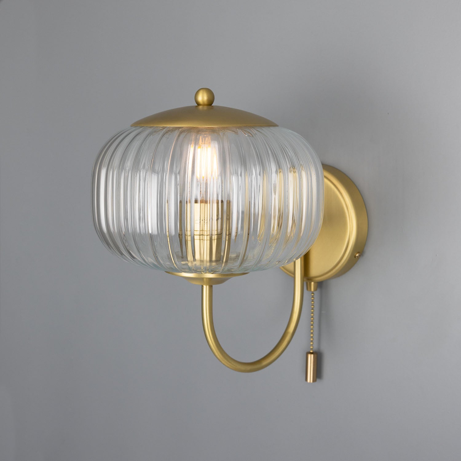 Wall-mounted light fixture with gold finish and clear glass shade on a gray background