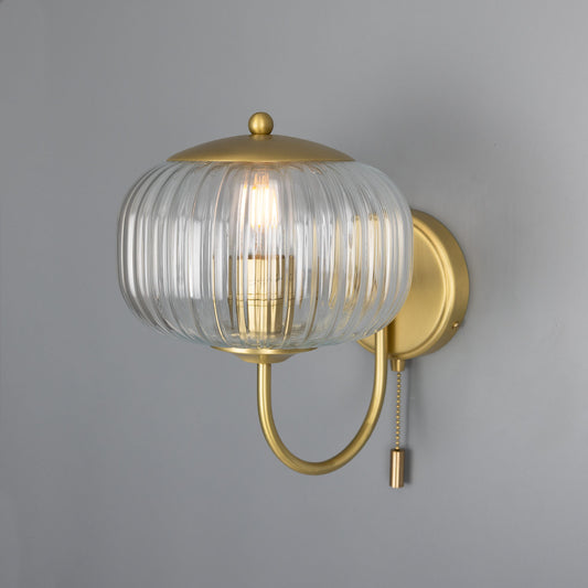 Wall-mounted light fixture with gold finish and clear glass shade on a gray background